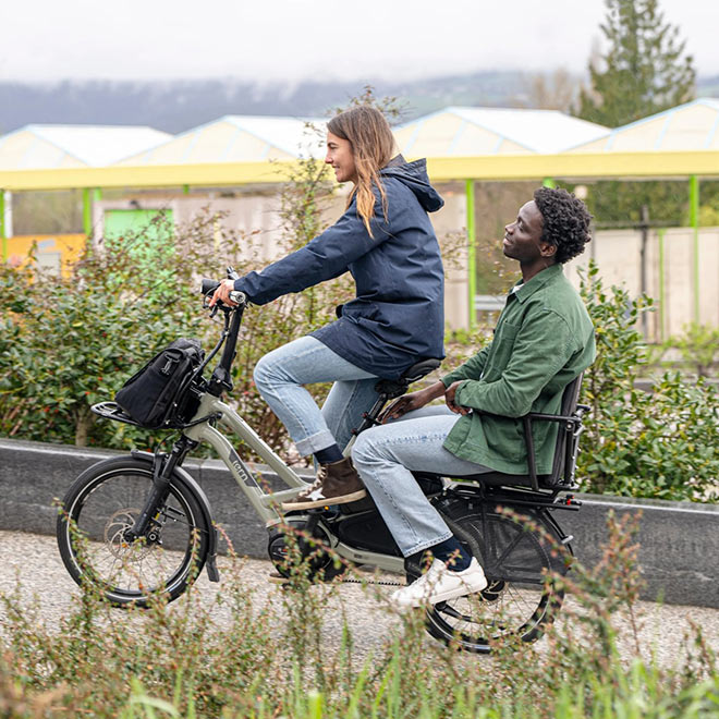 A woman riding a Tern HSD cargo e-bike with a man on the passenger seat and a bag on the front rack. 