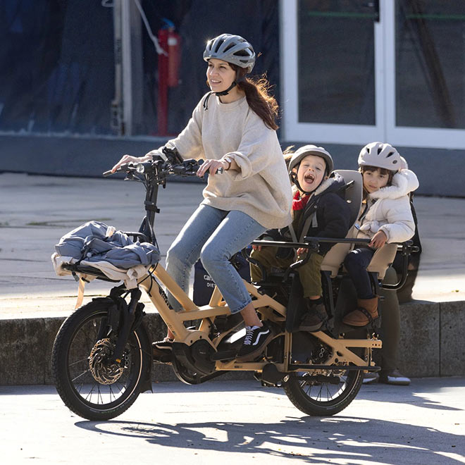 A mom riding a Tern GSD cargo e-bike with two young kids on the child seats and a bag strapped on the front rack. 