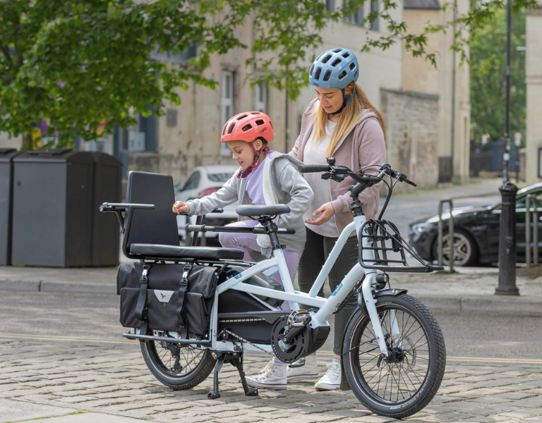 Adult and child with helmets standing beside a cargo bike on a city street.