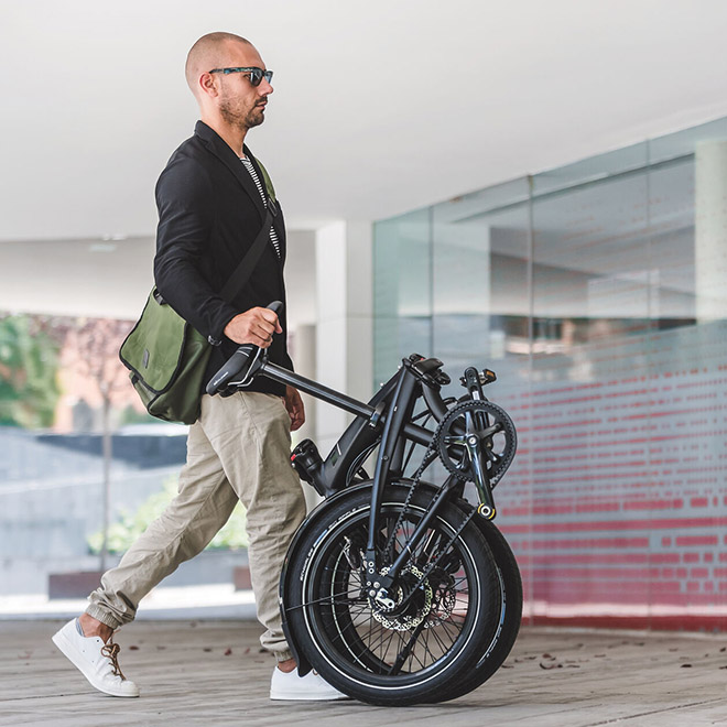 A man walks through an indoor space while rolling a folding bike by its seat post.