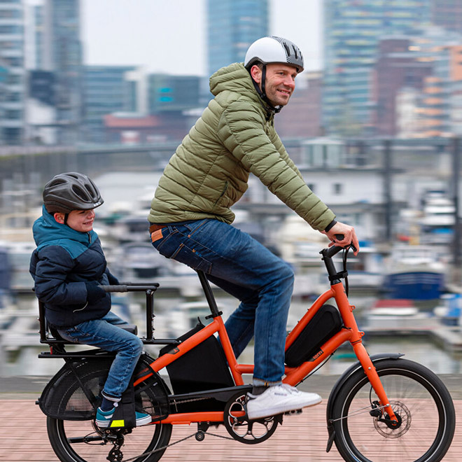 A man rides an orange short haul cargo bike with a child passenger through an urban area. 