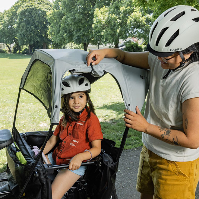 A child sits in a bike passenger seat under the Sun Shield Mini, while an adult adjusts the protective canopy in a park. 