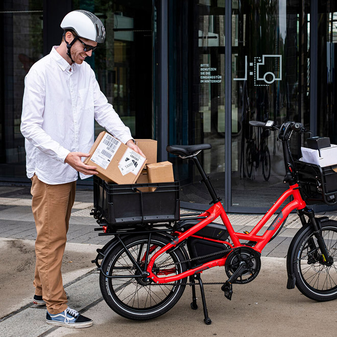 A man loads cardboard boxes onto a red HSD electric cargo bike equipped with the Cargo Tray outside a building.