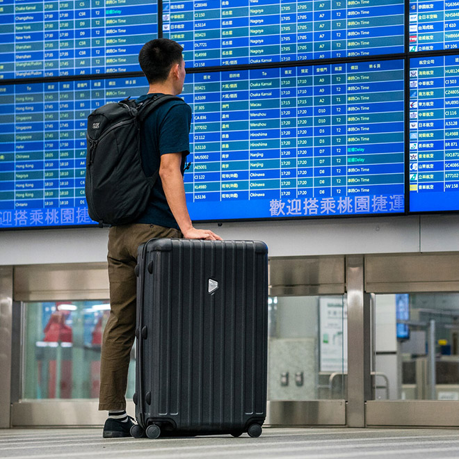 A man stands in an airport with a AirPorter Slim and backpack beneath flight information boards.