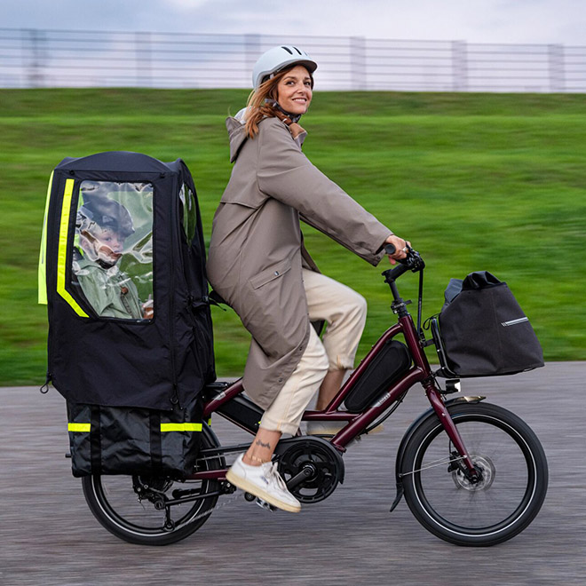 A woman rides a Quick Haul e-cargo bike carrying a child in a child seat equipped with a Rain Shield Mini.