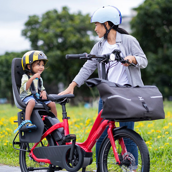 A woman rides a NBD, an ultra low-step ebike with a child in the rear seat and a bag attached to the front.