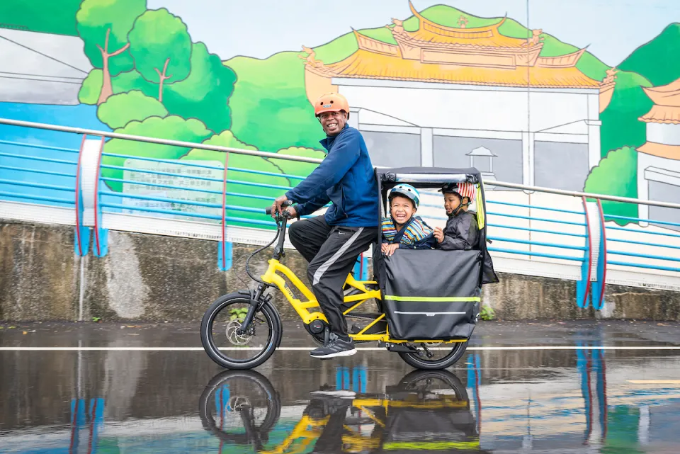 Family of three riding a GSD Gen 2 family electric bike, with the two kids sitting in the Clubhouse Fort weather canopy on the bike's rear rack.