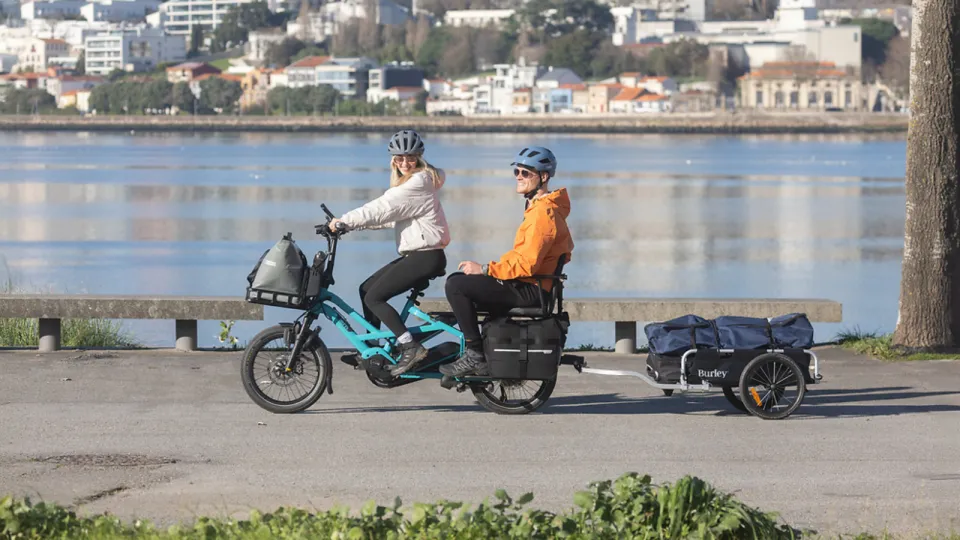 A woman riding a Tern GSD Gen 3 e-cargo bike, towing a trailer and with her partner in the passenger seat