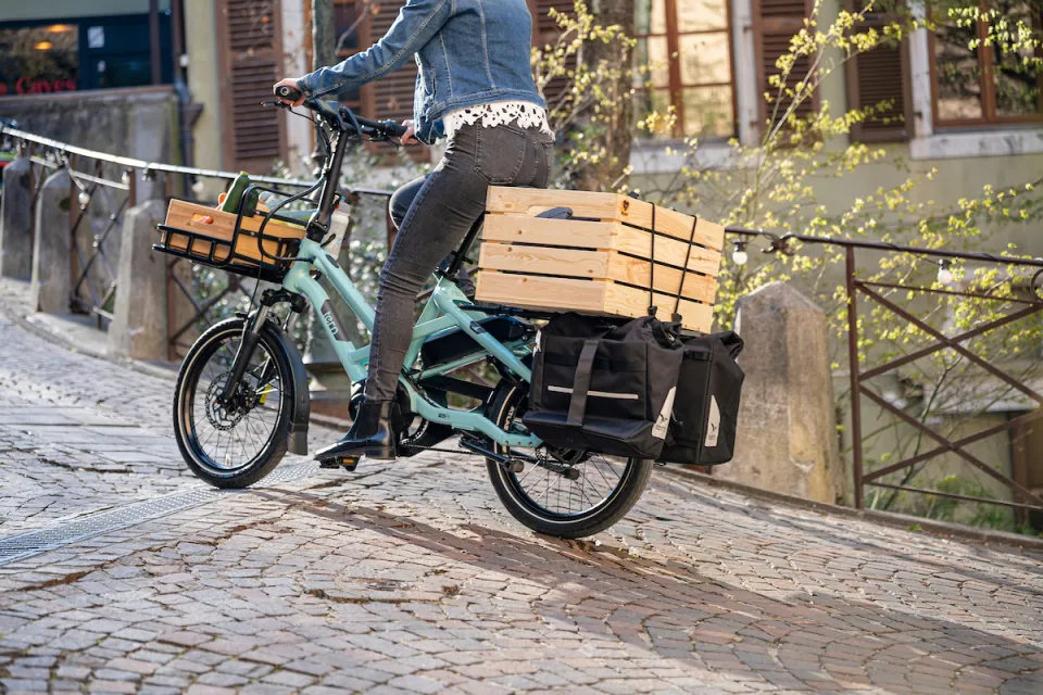 A woman riding a turquoise Tern HSD Gen 2 electric bicycle up a hill while carrying cargo on the bike's front and rear racks.
