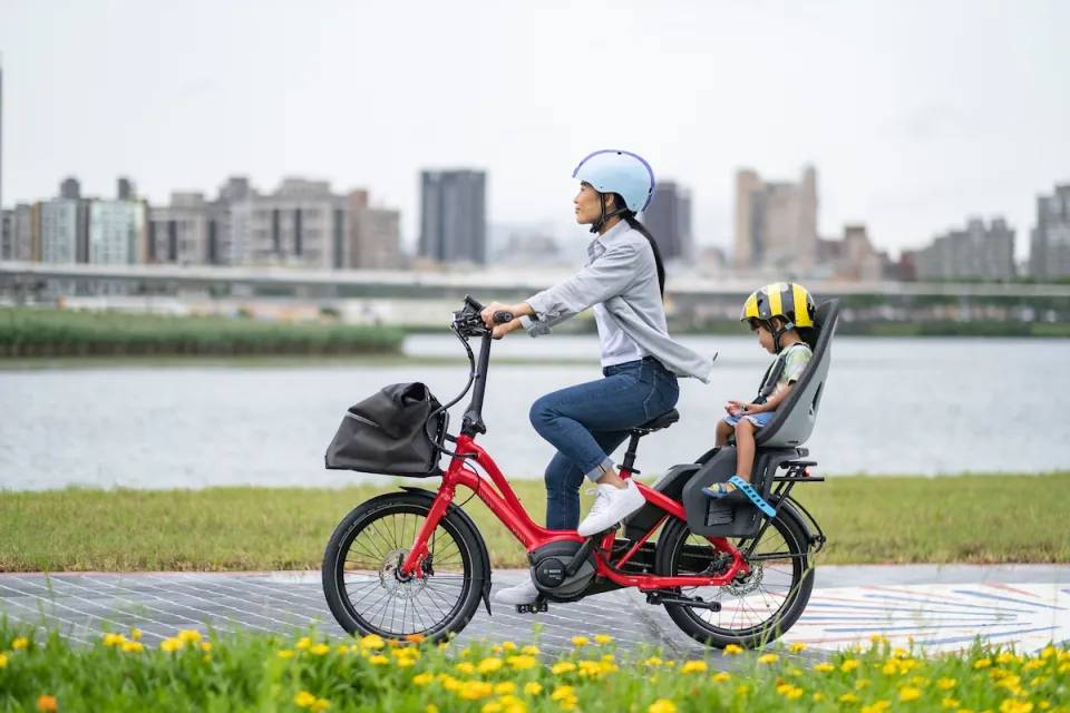 A mother riding a red Tern NBD electric bicycle while carrying her child, who's sitting in a child seat on the bike's rear rack.