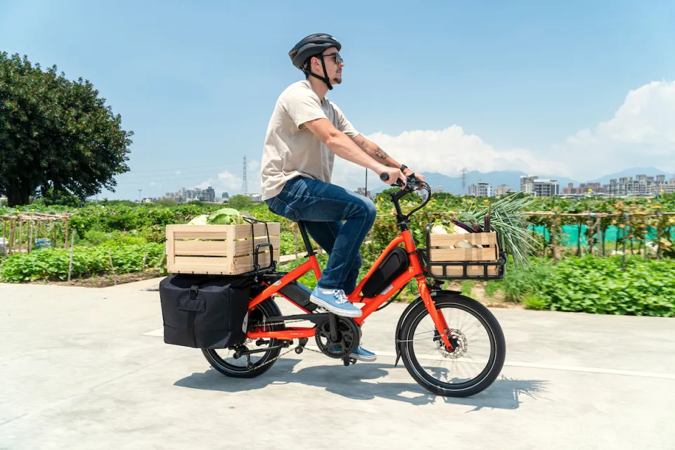 A man riding an orange Tern Quick Haul electric bicycle on a farm while carrying cargo on the bike's front and rear racks.