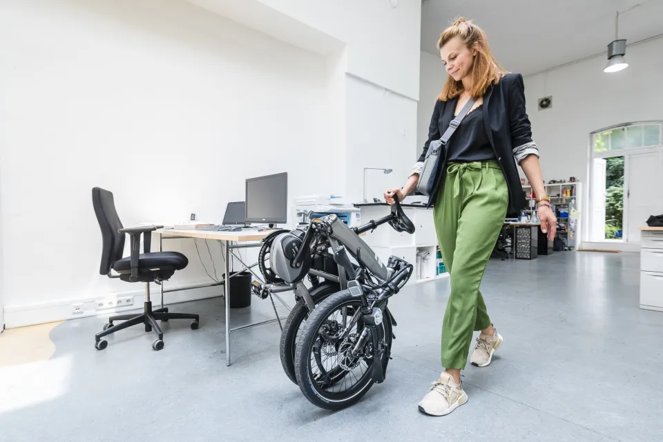 Silver Tern Vektron folding e-bike, folded and being rolled into an office by a woman.