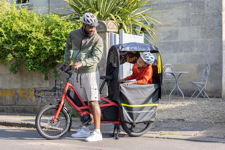 A father riding a red Tern Quick Haul Long electric bicycle, with his child sitting in a Clubhouse Fort rain canopy on the bike's rear rack.