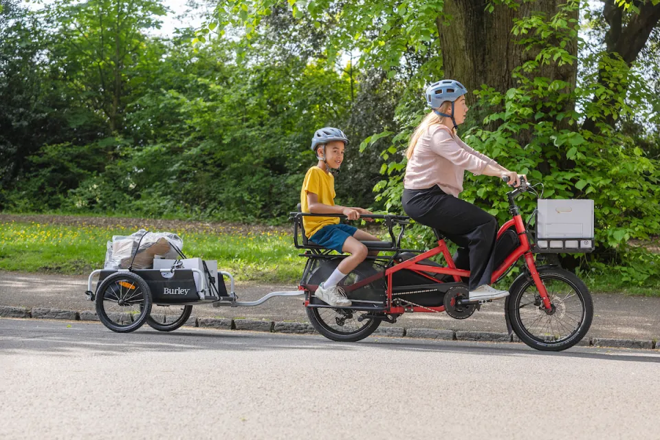 A woman riding a red Tern Quick Haul Long e-cargo bike, with her child in the rear rack's passenger seat and towing a trailer.