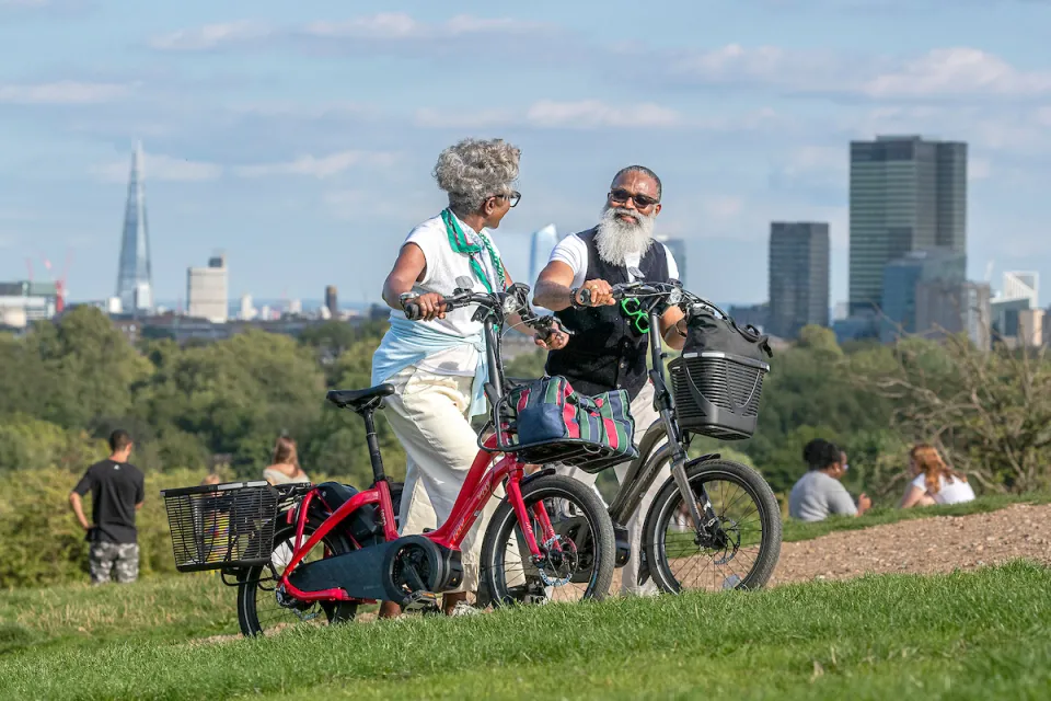 An elderly couple walking as they're pushing their red and silver Tern NBD electric bicycles up a hill in London, UK.