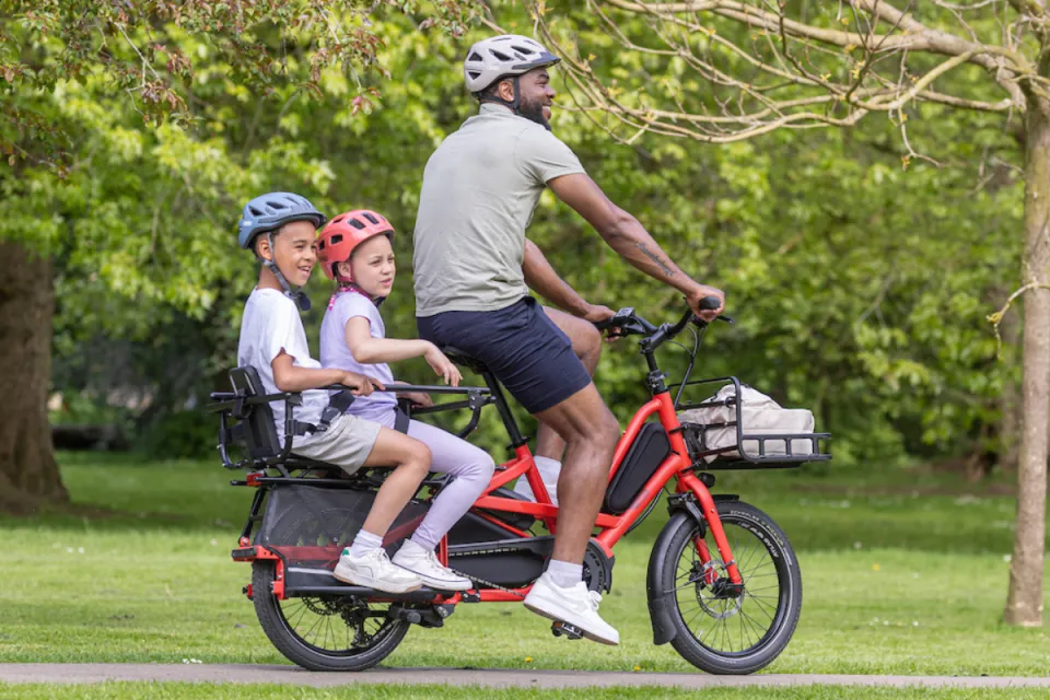 Family of three riding a Tern Quick Haul Long e cargo bike, with two kids sitting on the rear rack in the Clubhouse passenger rail.