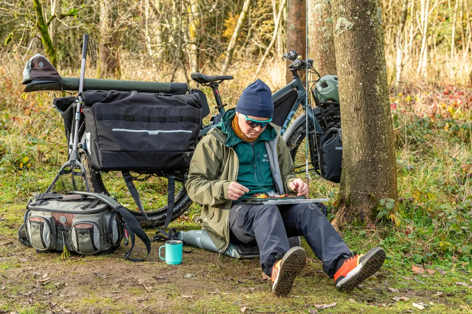 Man sitting under a tree in a forest with his Tern Orox electric adventure cargo bike and Cargo Hold 72 Panniers in the background, fishing gear beside him.