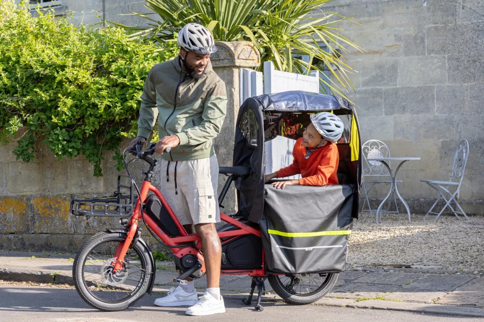 A dad riding a Tern Quick Haul Long cargo bike with his son on the back seat in a rain canopy