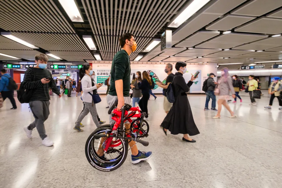 A man carrying a folded Tern BYB folding bike, walking across a busy metro station.