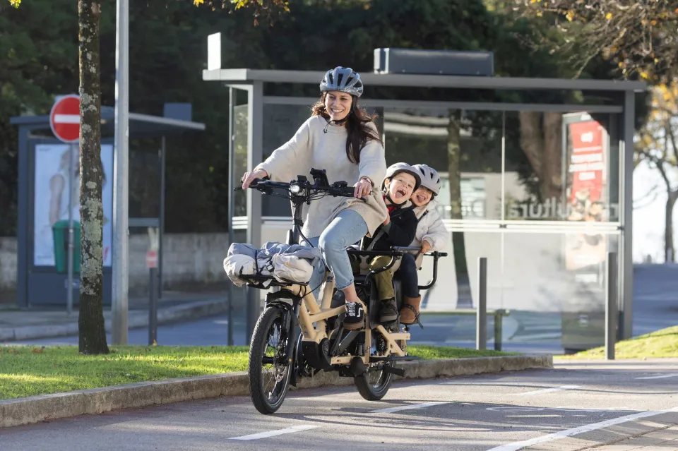 A mom riding a Tern GSD cargo e-bike with two children on the back, protected by the enclosed Clubhouse passenger rails.