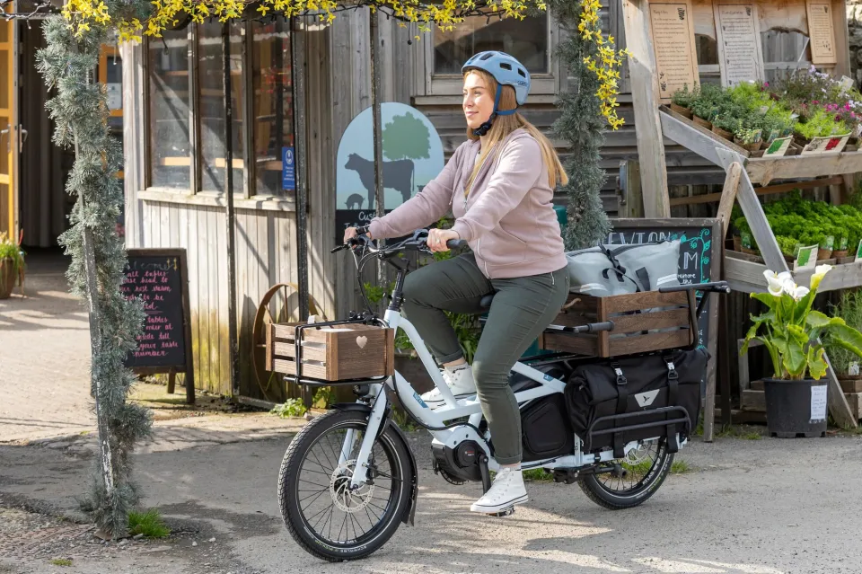 A woman riding a compact HSD longtail cargo bike with a crate on the back that contains a large carryall bag.